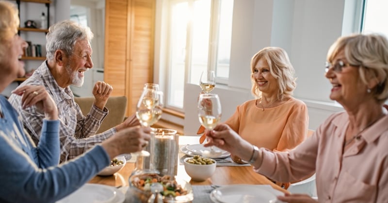 Four older adults smile and raise wine glasses around a dining table filled with food, sharing a warm, cheerful meal in a sunlit home.