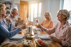 Four older adults smile and raise wine glasses around a dining table filled with food, sharing a warm, cheerful meal in a sunlit home.