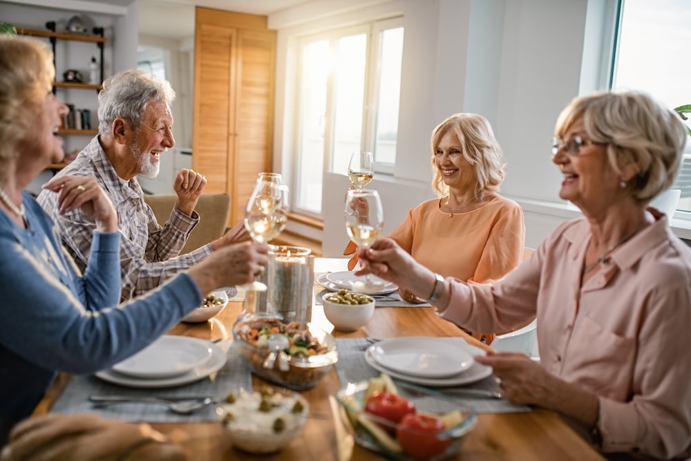 Four older adults smile and raise wine glasses around a dining table filled with food, sharing a warm, cheerful meal in a sunlit home.
