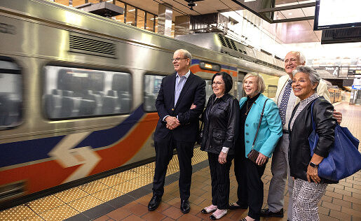 A group of five people stands smiling on a train platform as a train speeds by