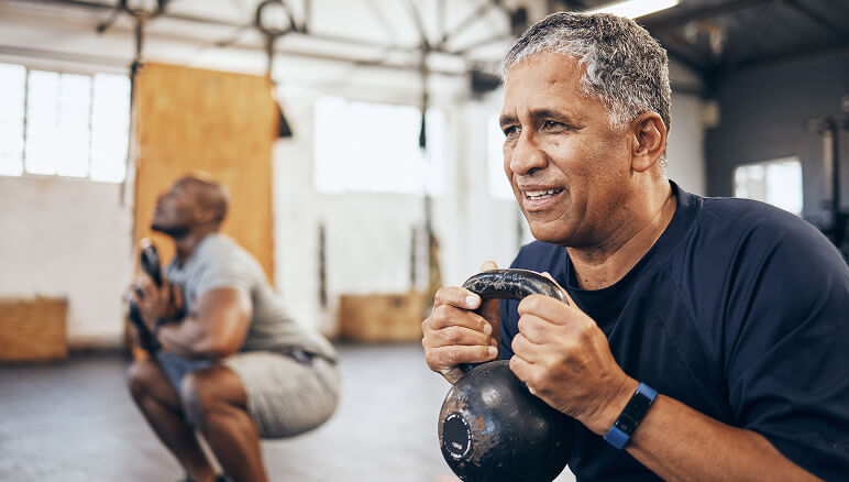 Man holding a kettlebell with determination in a gym, while another man performs squats in the background