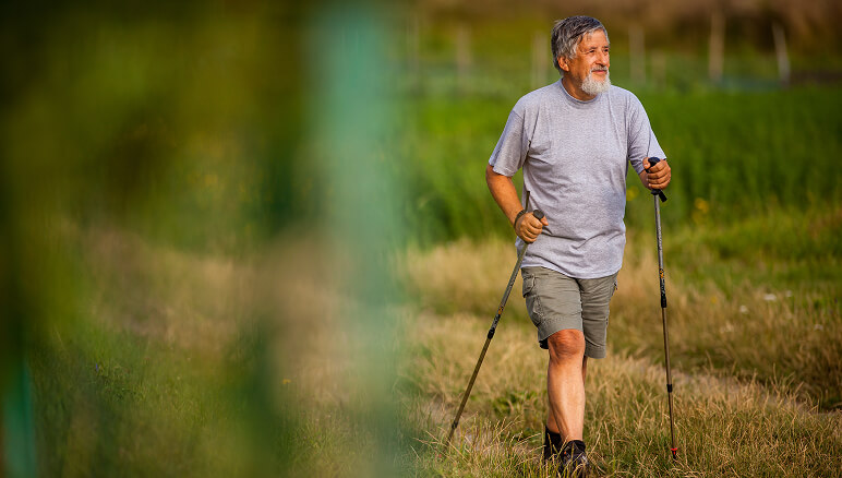 Man with trekking poles walks through lush green field, wearing a gray shirt and shorts
