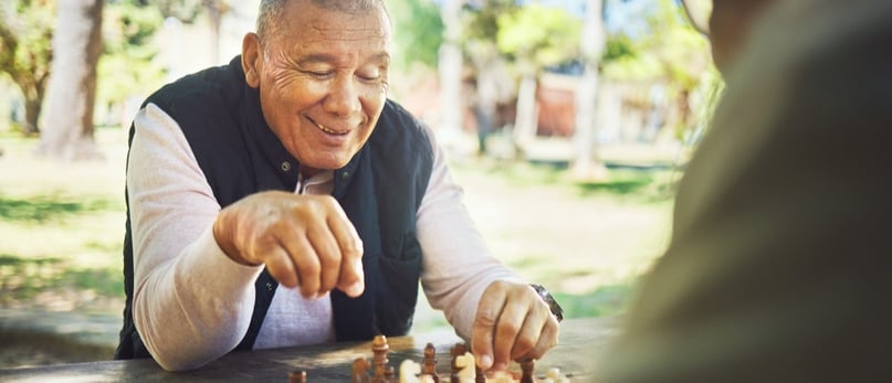 Man smiling while playing chess outdoors on a wooden table. 