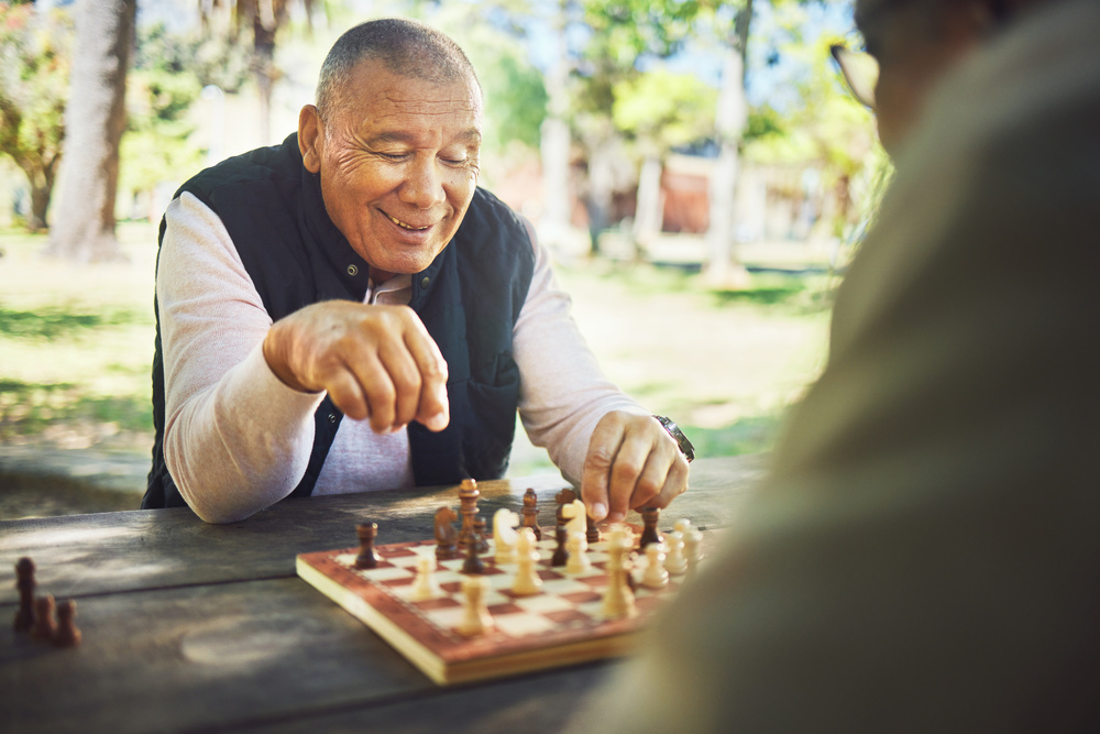Man smiling while playing chess outdoors on a wooden table. 