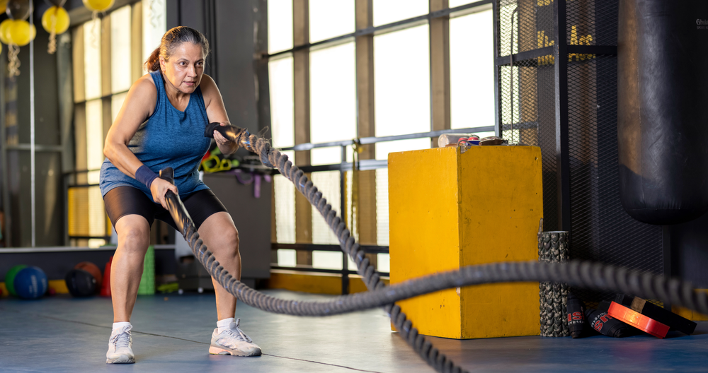 A woman in a blue tank top and black shorts performs a battle rope workout in a gym.